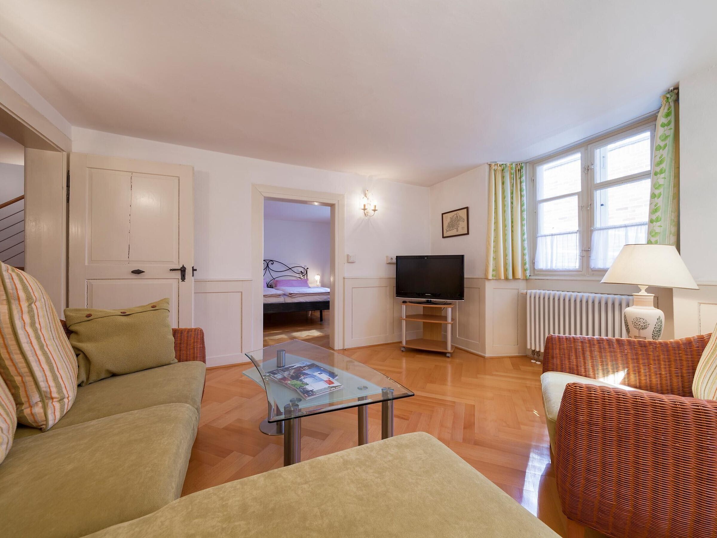 A living room with a sofa, armchair, glass coffee table, TV stand, and a bed in the background.
