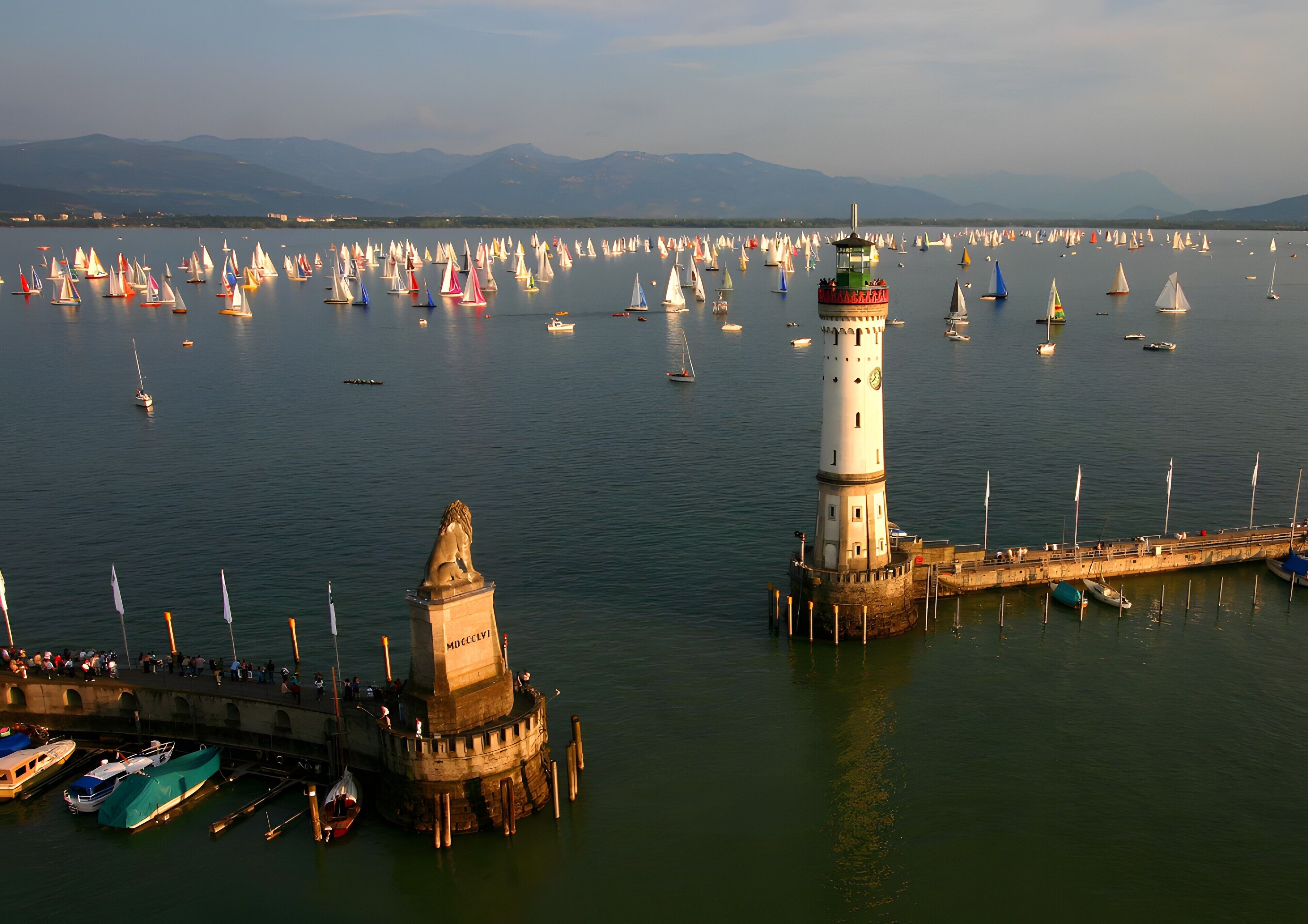 A lighthouse and a pier with boats docked, surrounded by a calm sea and distant mountains.