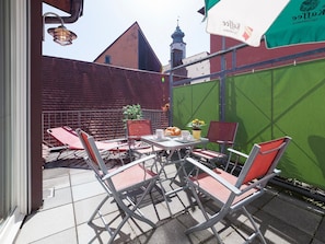 Un patio al aire libre con sillas y una mesa de metal, un muro verde y un edificio con techo rojo al fondo.