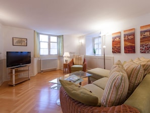 Un salon comprenant un téléviseur, une table basse en verre, un canapé, un fauteuil en osier et un radiateur.