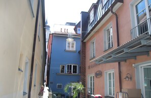 A narrow street with buildings, including a blue building with a window and a door, and a pink building with a balcony.