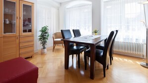 A dining area with a wooden table, chairs, a red bench, a cabinet with glass doors, a potted plant, and large windows with sheer curtains. - Apartment 'Kaufmannshaus' with Wi-Fi (Lindau am Bodensee)