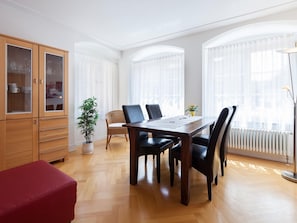 A dining area with a wooden table, chairs, a red bench, a cabinet with glass doors, a potted plant, and large windows with sheer curtains.