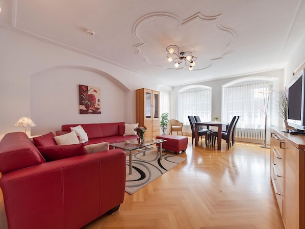 A modern living room with a red sofa, a glass coffee table, a dining area with a table and chairs, and a television on a wooden cabinet. - Apartment 'Kaufmannshaus' with Wi-Fi (Lindau am Bodensee)