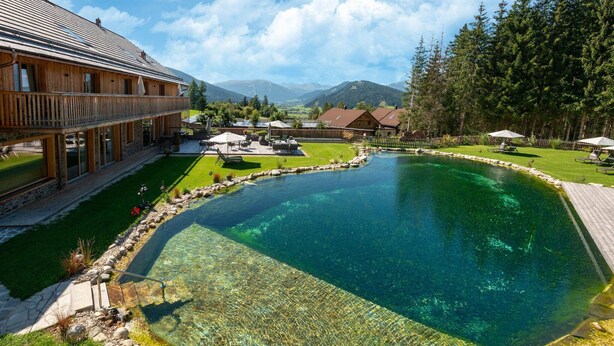 A large wooden building with a swimming pool, surrounded by a well-maintained lawn and trees.