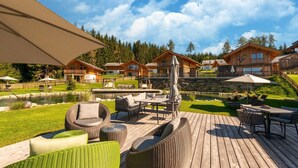 A wooden deck with outdoor furniture, a pond, and a row of wooden cabins in the background.
