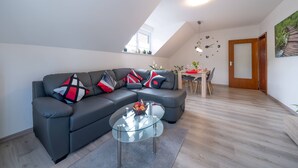 Un salon moderne avec un canapé d’angle, une table basse en verre, un coin repas avec une table en bois et une horloge au mur.