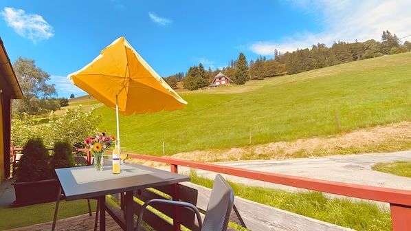 Ein Tisch mit einem gelben Sonnenschirm, einer Flasche und einem Blumenstrauß auf einem Balkon mit Blick auf ein grünes Feld und ein Haus im Hintergrund.