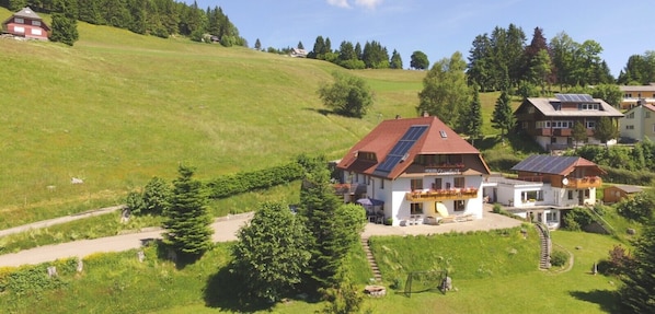 Una propiedad en una ladera con un edificio principal de techo inclinado y paneles solares, rodeada de abundante vegetación y un sendero sinuoso.