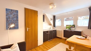 A compact kitchen with a wooden door, a dining table with a yellow tablecloth, a TV on a stand, and a window with curtains. - Pension Elisabeth Apartment No. 11 with Mountain and Village View (Todtnau)