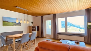 Un salon moderne avec un plafond en bois, un coin repas avec une table et des chaises, un canapé, une grande fenêtre donnant sur l’extérieur et un téléviseur fixé au mur.