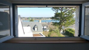 A window view of a residential area with houses, a tree, and a body of water. - Croix du Sud - cosy town centre house (La Trinité-sur-Mer)