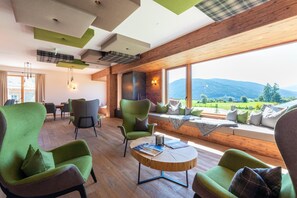 Um interior moderno com cadeiras estofadas em verde, uma mesa de centro redonda de madeira e uma grande janela com vista para uma paisagem montanhosa.