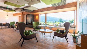 A modern interior with a wooden floor, green armchairs, a round coffee table, and a large window offering a mountain view.