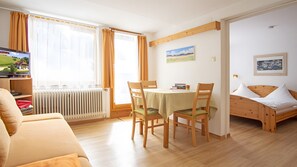Een kamer met houten vloer, een eettafel met stoelen, een bed, een radiator, een televisie en een raam met gordijnen.