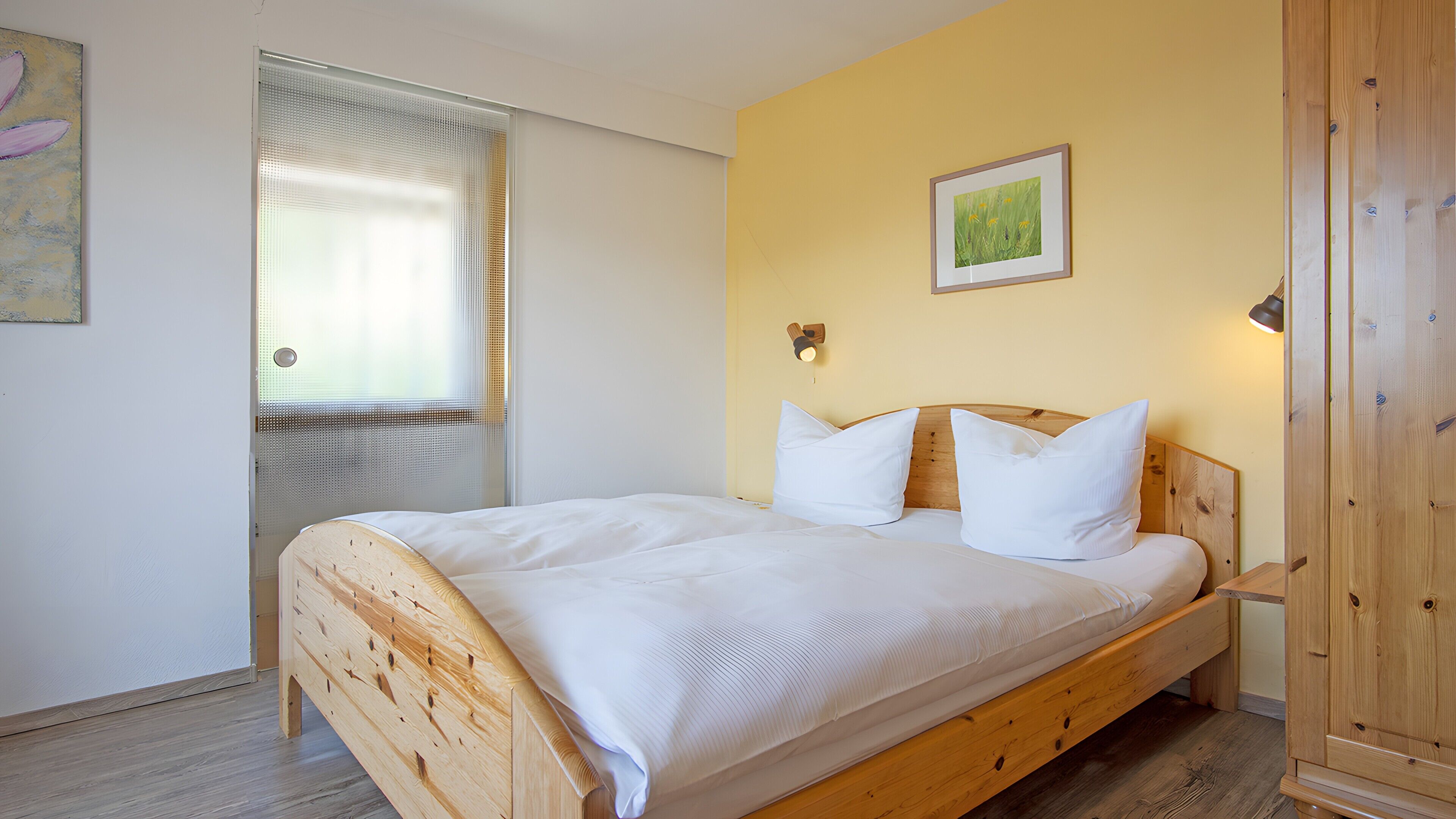 A wooden bed with white bedding, a wooden wardrobe, and a wall-mounted light fixture.