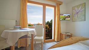 Ein Hotelzimmer mit einem Bett, einem Tisch mit Teller und Buch, einem Sessel, einem Fernseher an der Wand und einem Balkon mit Blumen.