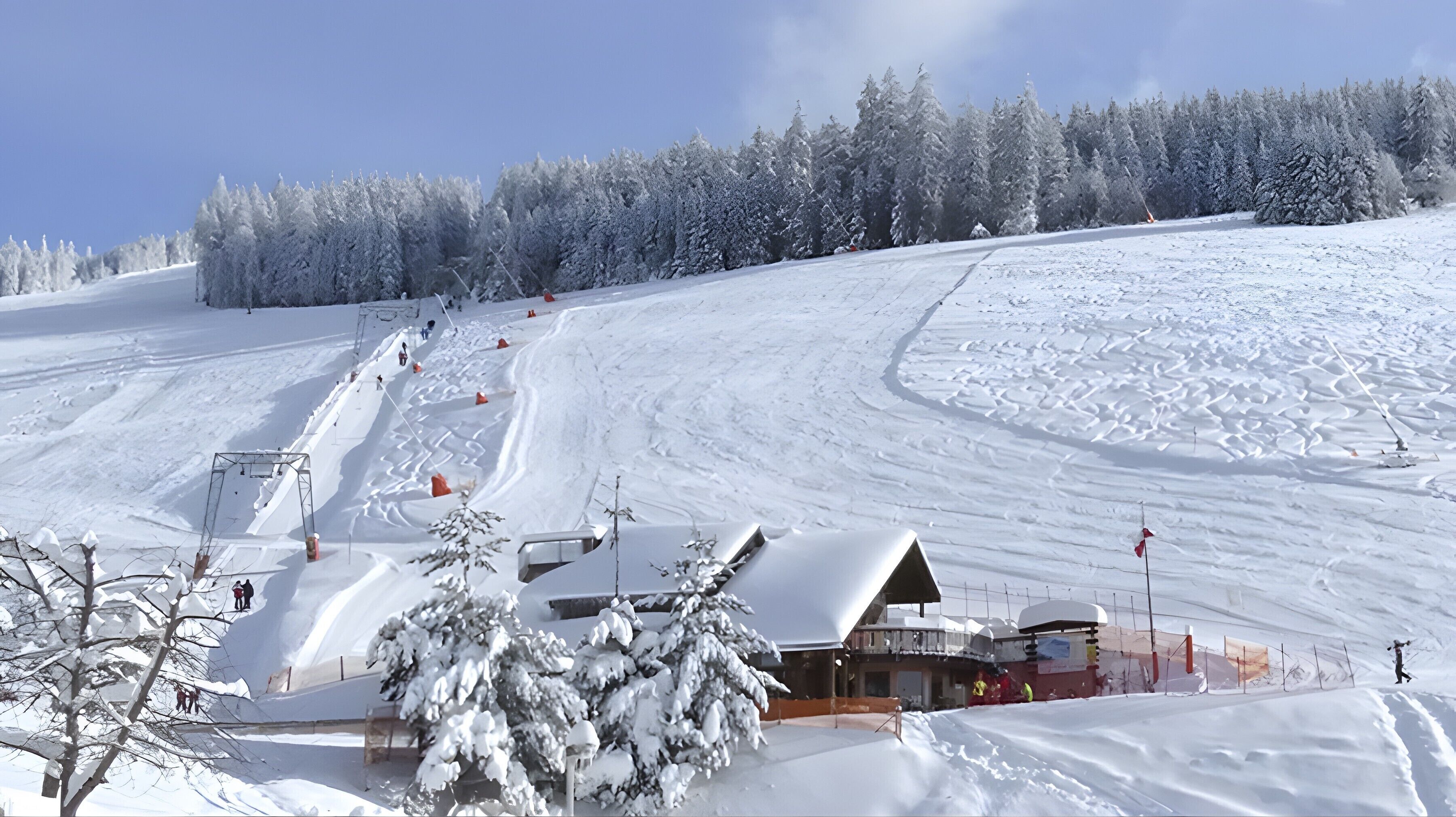 Une piste de ski enneigée, avec un chalet, des arbres et un ciel dégagé.