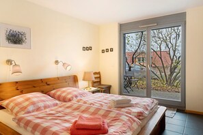 Une chambre avec une tête de lit en bois, deux lits avec une literie à carreaux roses, une table de chevet avec une lampe, une chaise et une grande fenêtre donnant sur des arbres et un immeuble.
