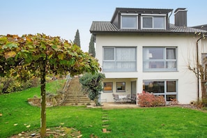 Una casa de dos pisos con jardín, un patio con mobiliario exterior y una escalera que lleva a la entrada.