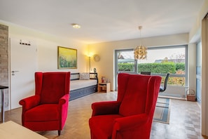 Una habitación con una cama, dos sillones rojos, una ventana con vistas, una lámpara y un reloj en la pared.
