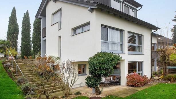 Ein modernes, zweistöckiges Haus mit flachem Dach, großen Fenstern und einem gepflegten Garten.