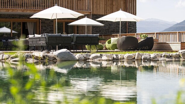A pool with lounge chairs and umbrellas, a stone building with wooden slats, and a mountainous backdrop.