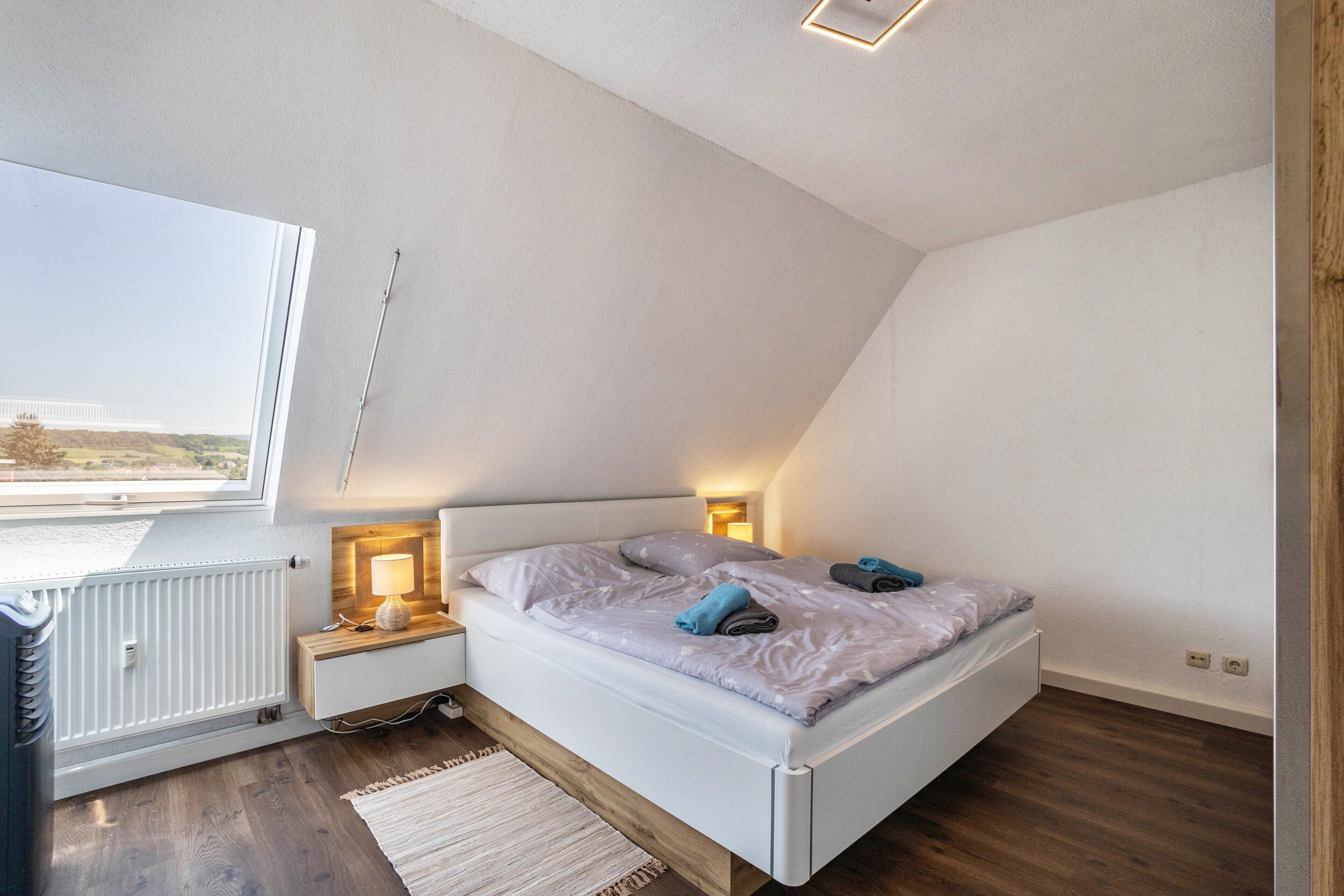 Ein Schlafzimmer mit schräg stehender Decke, einem Bett mit Kissen, Nachttischlampen, einem Heizkörper und Blick durch ein Fenster.