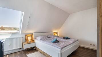Ein Schlafzimmer mit schräg stehender Decke, einem Bett mit Kissen, Nachttischlampen, einem Heizkörper und Blick durch ein Fenster.
