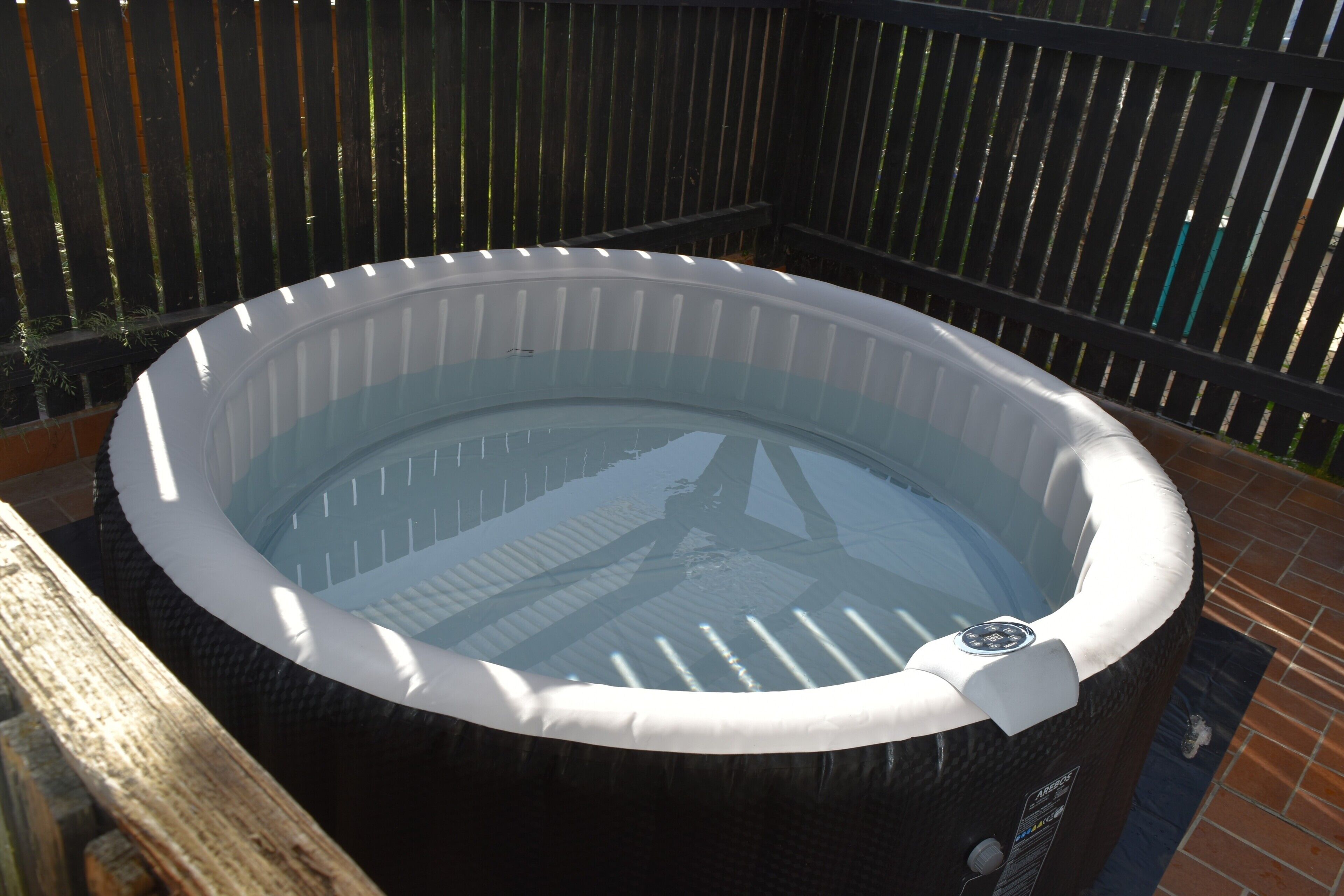 A hot tub with a control panel, surrounded by a wooden fence and a brick floor.