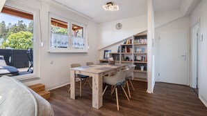 A room with a wooden dining table, chairs, a bookshelf, and a window with a view of trees.