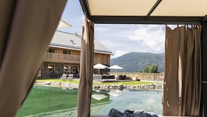 A room with a view of a swimming pool, outdoor furniture, and a wooden building in the background. - Chalet in Wölting with natural swimming pond (Wölting)