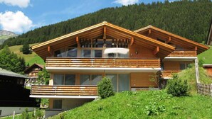 Een moderne houten chalet met balkon, grote ramen en een bergachtige achtergrond.
