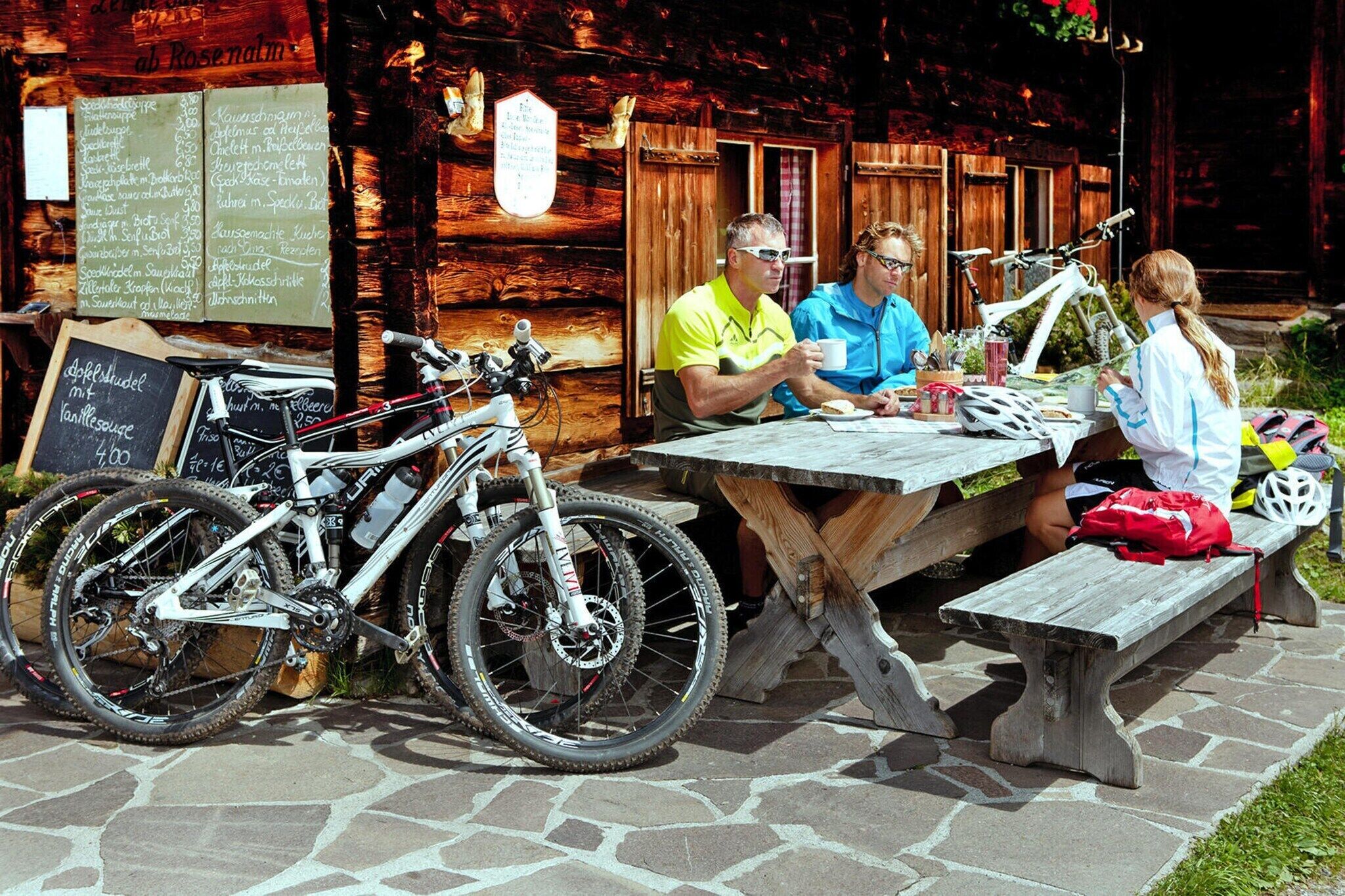 Ein hölzerner Außenbereich zum Sitzen mit Tisch und Bänken, in der Nähe sind Fahrräder abgestellt und ein Speiseplan ist im Hintergrund sichtbar.