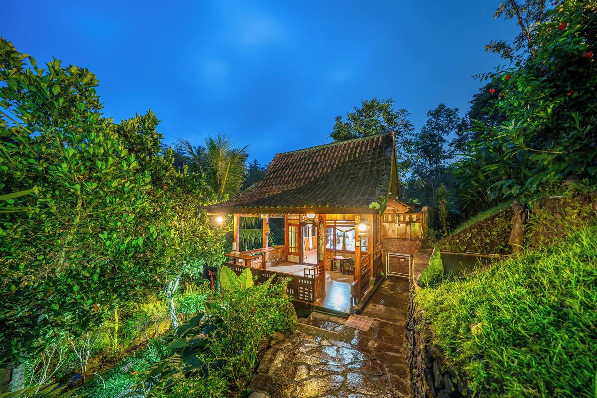 A traditional wooden house with a tiled roof, surrounded by lush greenery and a stone pathway leading to the entrance.