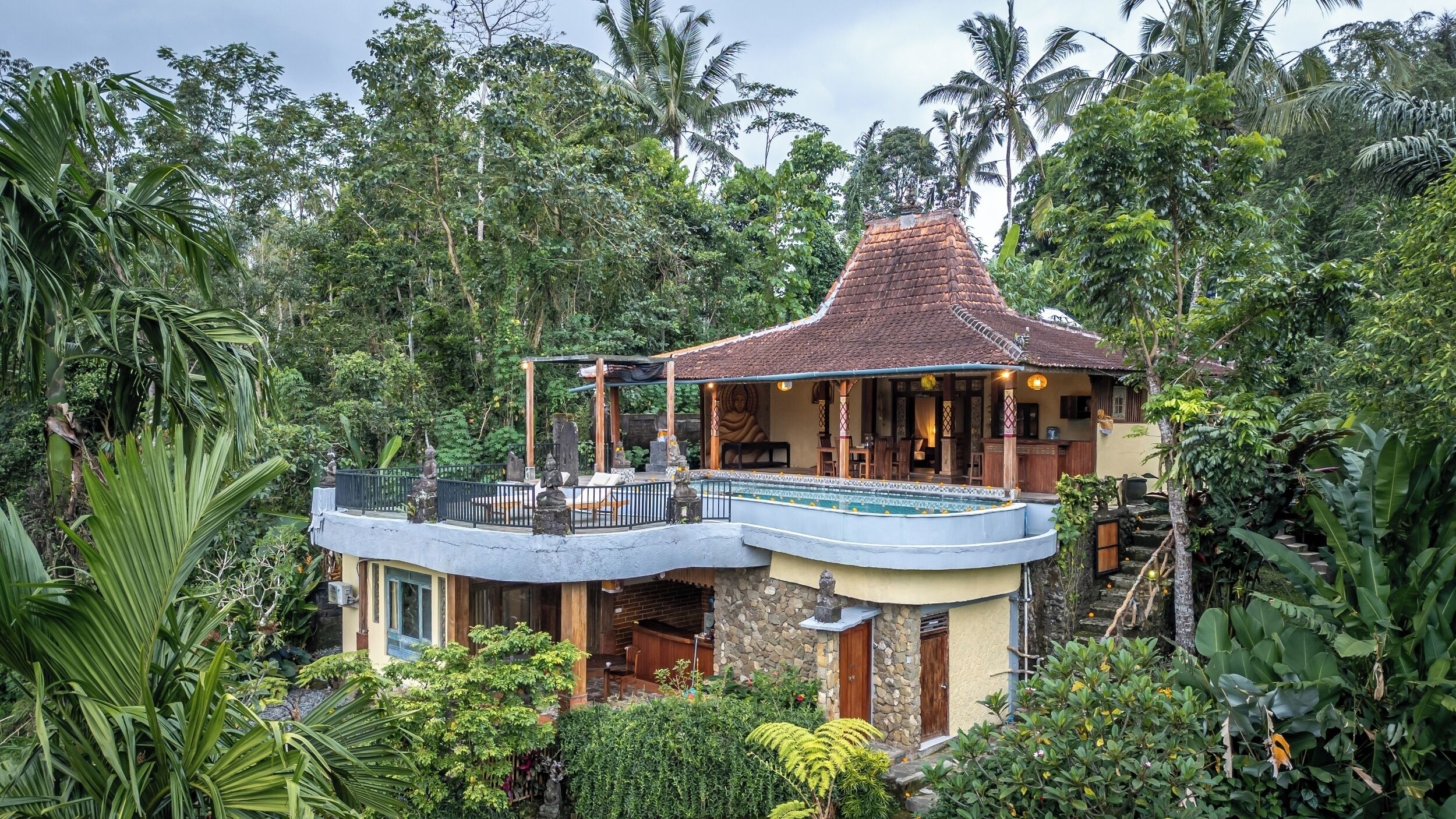 A two-story house with a balcony, surrounded by lush tropical vegetation.