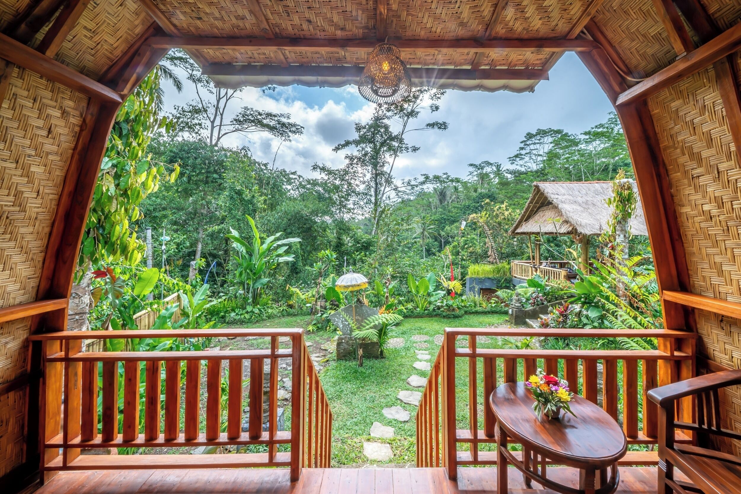 Una terraza de madera con una mesa y sillas, con vistas a un jardín exuberante y una estructura con techo de paja al fondo.