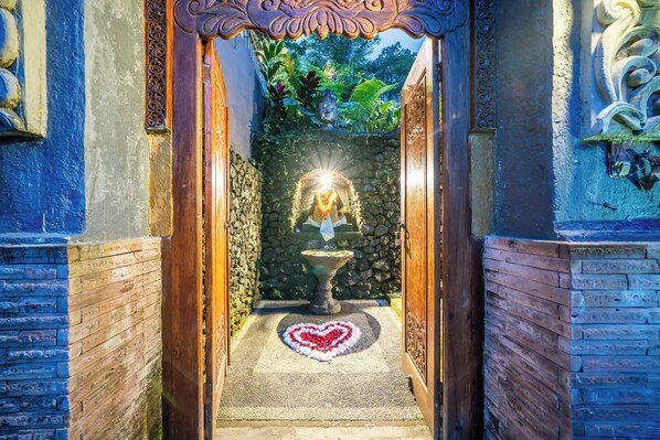 A narrow pathway leading to a stone wall with a statue, surrounded by wooden panels and a heart-shaped decoration on the floor.