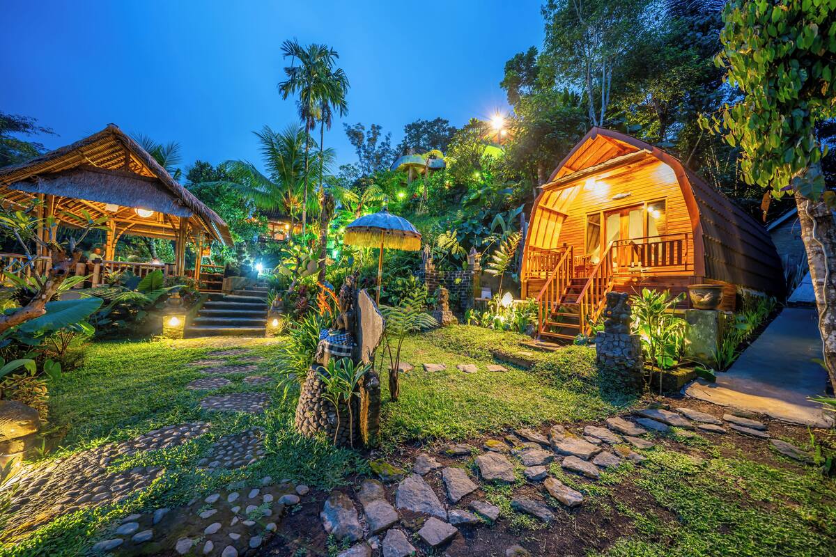 A well-lit, tropical resort with thatched-roof cabins, a stone pathway, and lush greenery.