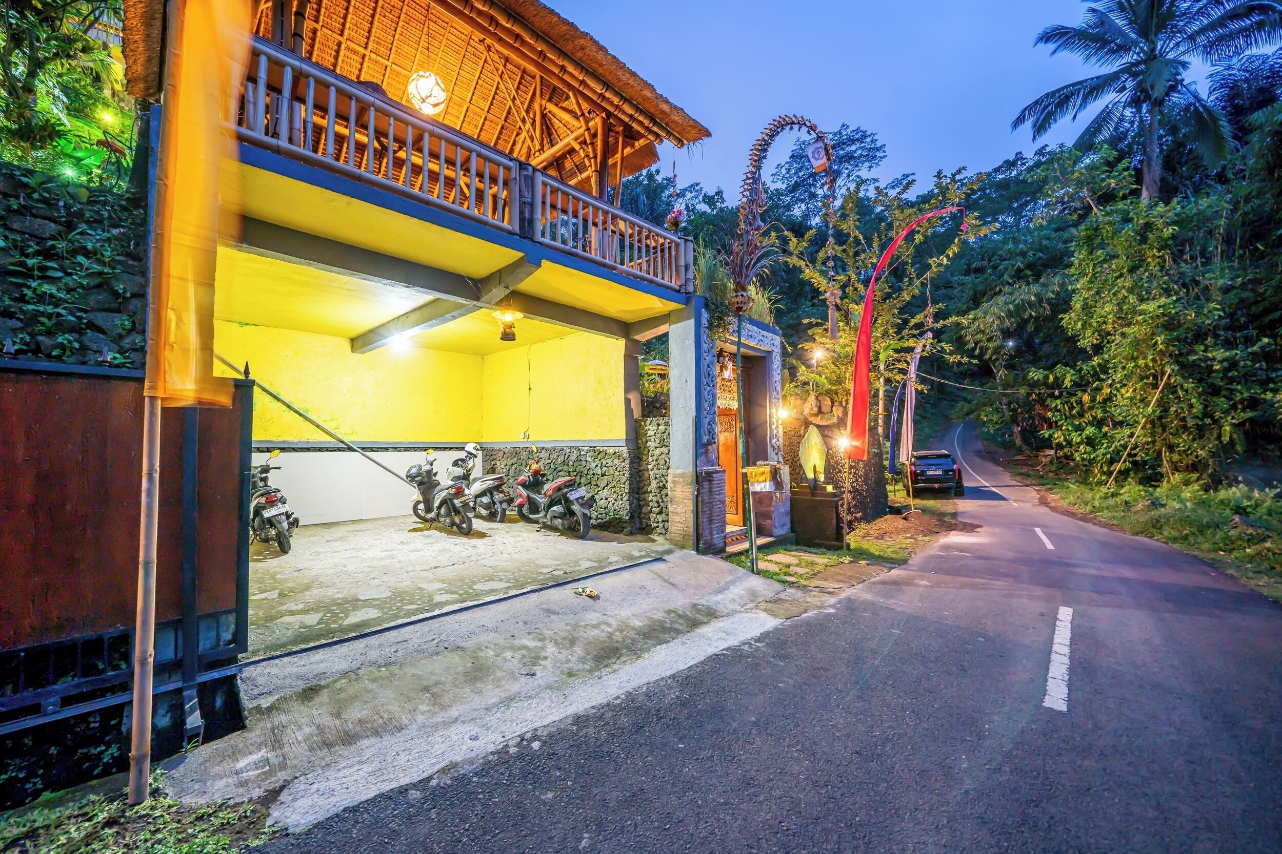 A two-story building with a balcony, a paved area with parked scooters, and a road with a curve.