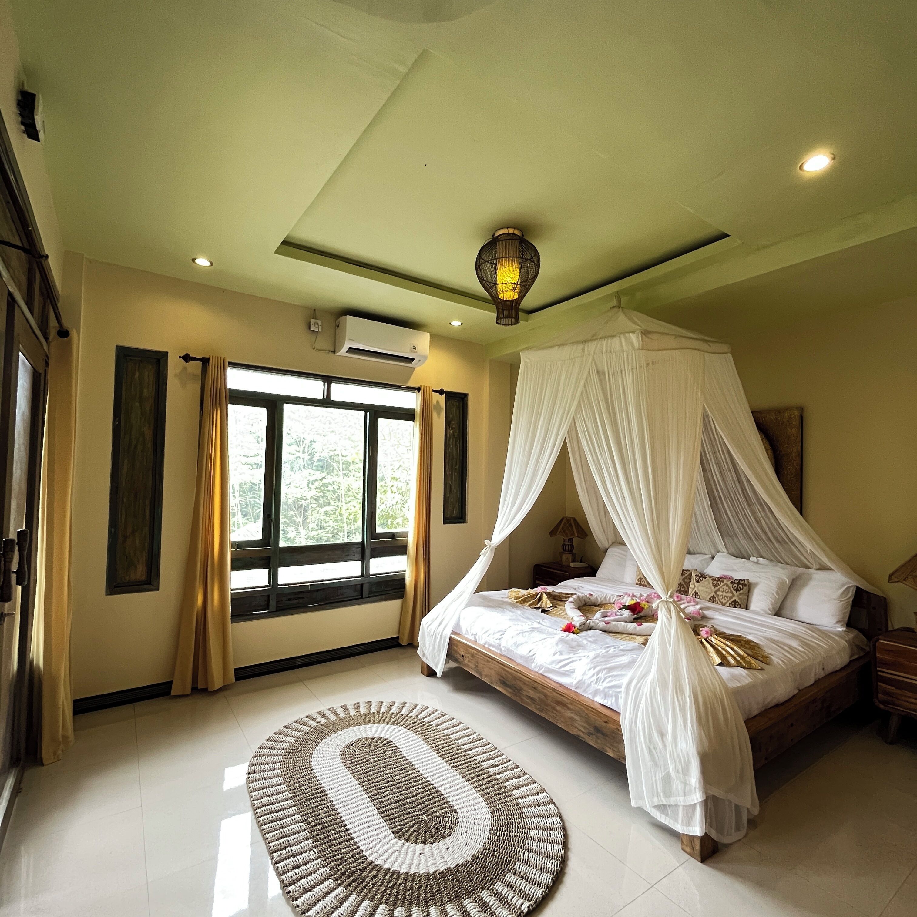 Een slaapkamer met een hemelbed, een groot raam met gordijnen, een airconditioning en een decoratief vloerkleed.