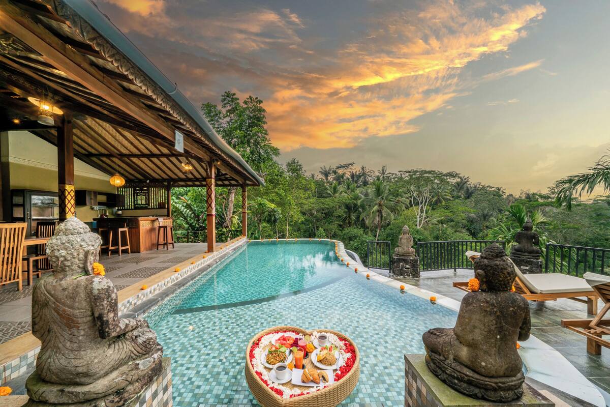 A serene pool area with a Buddha statue, a heart-shaped arrangement of food, and a bar area with outdoor seating.