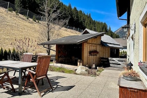 A wooden building with a sloped roof, a patio with chairs and a table, and a mountainous backdrop. - Alpine Holiday Home in Lungau (Vordermuhr)