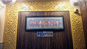 A reception area with a framed artwork above a 'RECEPTION' sign, decorative panels, and two standing fans. - Puri Inn (Varanasi)