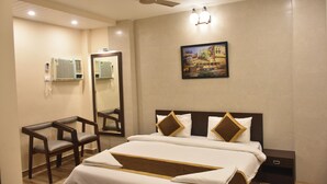 A hotel room with a bed, two chairs, a ceiling fan, a painting, and a nightstand. - Puri Inn (Varanasi)
