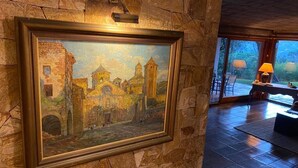 A framed painting of a historical building on a stone wall, a stone fireplace, and a view of a garden through large windows.