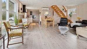 Ampla sala de estar com escada de madeira, mesa de jantar e cozinha ao fundo.