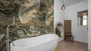 A modern bathroom with a freestanding bathtub, a marble wall, a wooden bench, and a window with blinds. - Agkyra Studio in port of Symi (Simi)