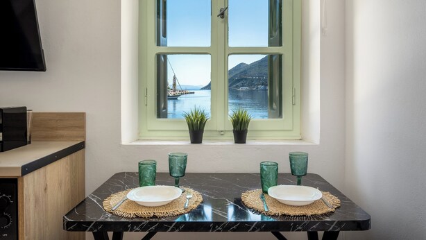 A dining table with two plates and green glasses, set against a window with a view of a body of water and mountains.