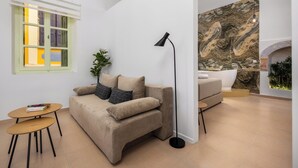 A modern living room with a beige sofa, a small wooden table, a floor lamp, and a large wall mural. - Agkyra Studio in port of Symi (Simi)
